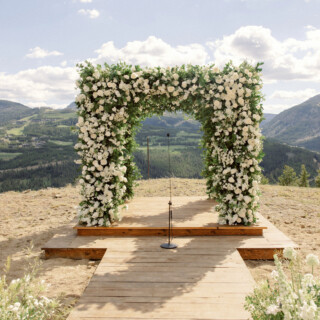 Scenic mountaintop wedding ceremony with white floral arch and panoramic mountain views in Big Sky Montana.