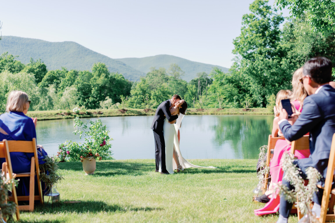 Bride and groom kiss by a scenic pond during a Vermont countryside outdoor wedding ceremony.