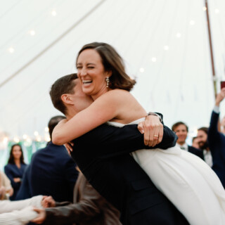 Bride laughing as groom lifts her during joyful wedding reception under glowing tent lights.