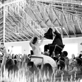 Bride and groom lifted during joyful Hora dance at rustic Thompson Island wedding celebration.