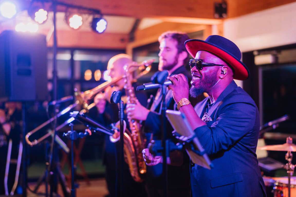 Male singer performing with live brass band under warm colorful lighting at indoor event.
