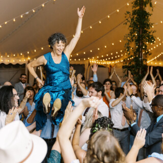 Bride lifted on chair during joyful wedding celebration under glowing tent lights in Bristol, Rhode Island.