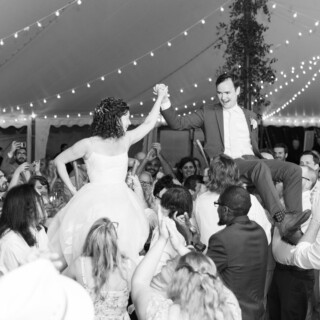 Bride and groom lifted on chairs during joyful outdoor wedding celebration under string lights.