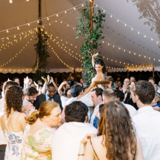 Bride lifted on chair during joyful Jewish wedding dance under glowing tent lights.