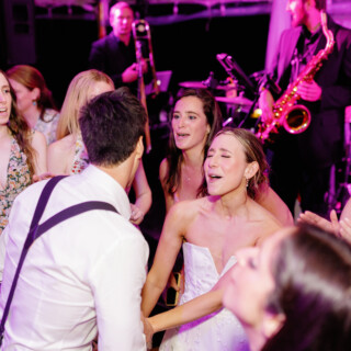 Bride and groom dancing joyfully with guests under vibrant pink and purple wedding lights.