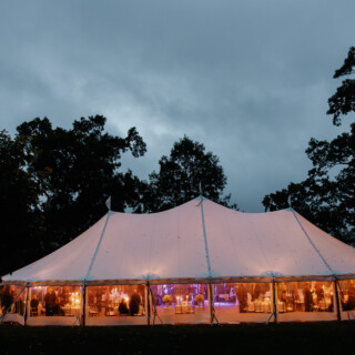 Glowing woodland wedding tent at dusk with warm lights and guests celebrating in Massachusetts.