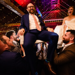 Bride and groom lifted during joyful Hora dance at rustic barn wedding celebration.