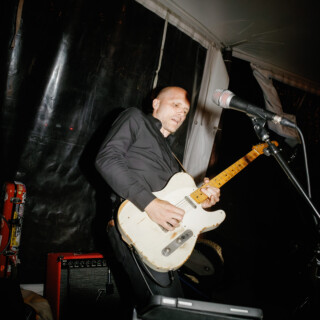 Bald guitarist performing live with vintage electric guitar under warm stage lights.