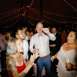 Guests joyfully dancing and singing under warm string lights at a Moraine Farm wedding reception.