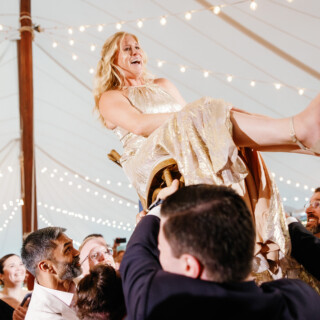 Bride in gold gown lifted on chair during joyful Moraine Farm wedding celebration.