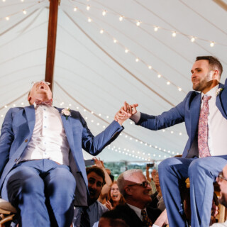 Two grooms lifted on chairs during joyful wedding celebration under glowing tent at Moraine Farm.