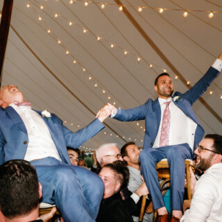 Two grooms lifted on chairs during joyful same-sex wedding celebration at Moraine Farm, Beverly MA.