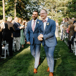 Two grooms walk hand in hand down a sunny outdoor aisle at Moraine Farm wedding.
