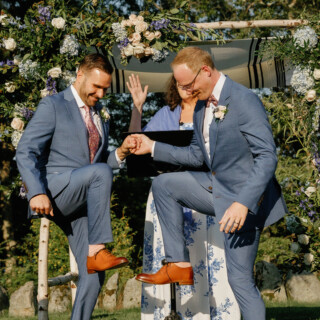 Two grooms joyfully break the glass under a floral chuppah at their outdoor Jewish wedding.