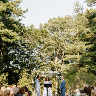 Two grooms exchange vows under a floral arch at an outdoor forest wedding ceremony.