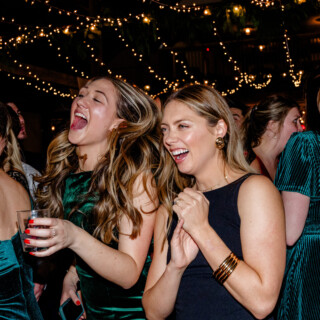 Two women laughing under glowing string lights at an elegant Vermont wedding celebration.