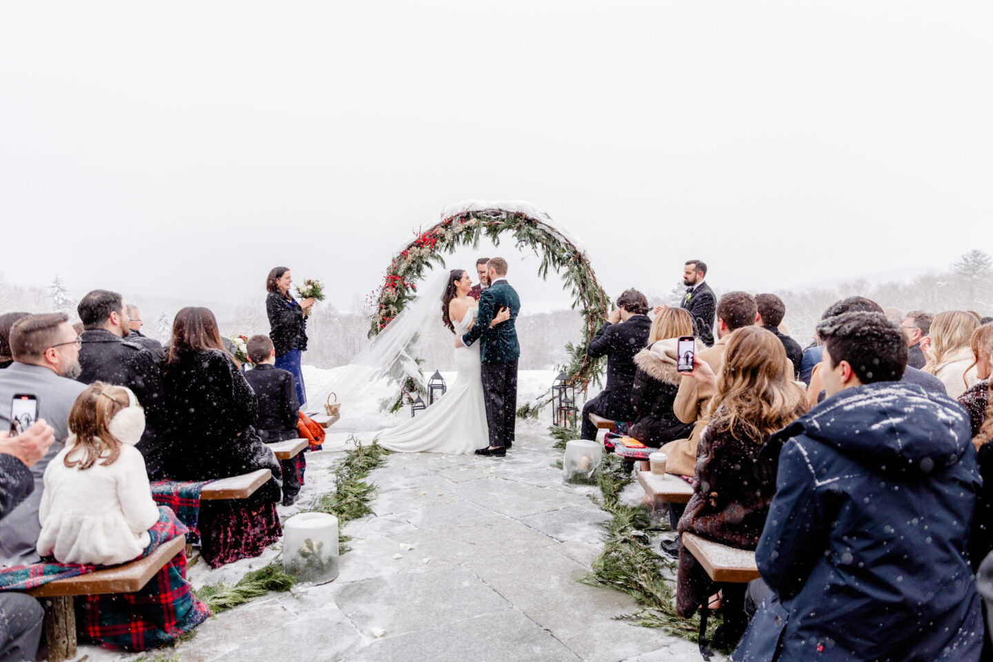 Bride and groom kiss under snowy evergreen arch at Mountain Top Inn winter wedding in Vermont.