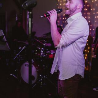 Male singer performing live under warm purple lights with band and glowing fairy light backdrop.