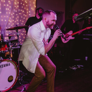 Male singer performing passionately with live band under warm, colorful stage lights.
