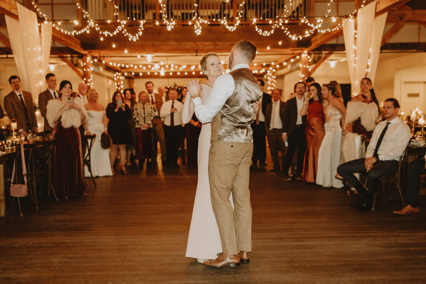 Rustic barn wedding reception with guests dancing under warm string lights and wooden beams.