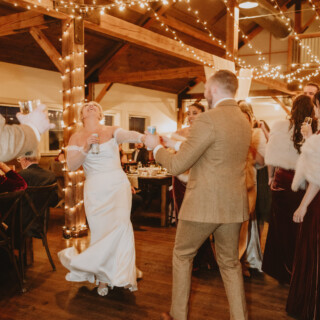 Bride and groom dancing joyfully at rustic Vermont barn wedding reception under warm string lights.