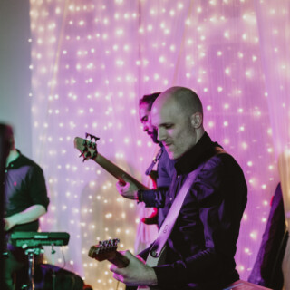 Bald guitarist performing live with band under warm pink and golden fairy lights at wedding.
