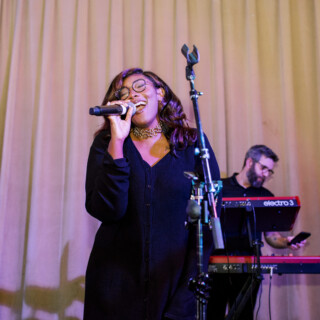 Passionate female singer performing live on stage with band under warm lighting and beige curtains.